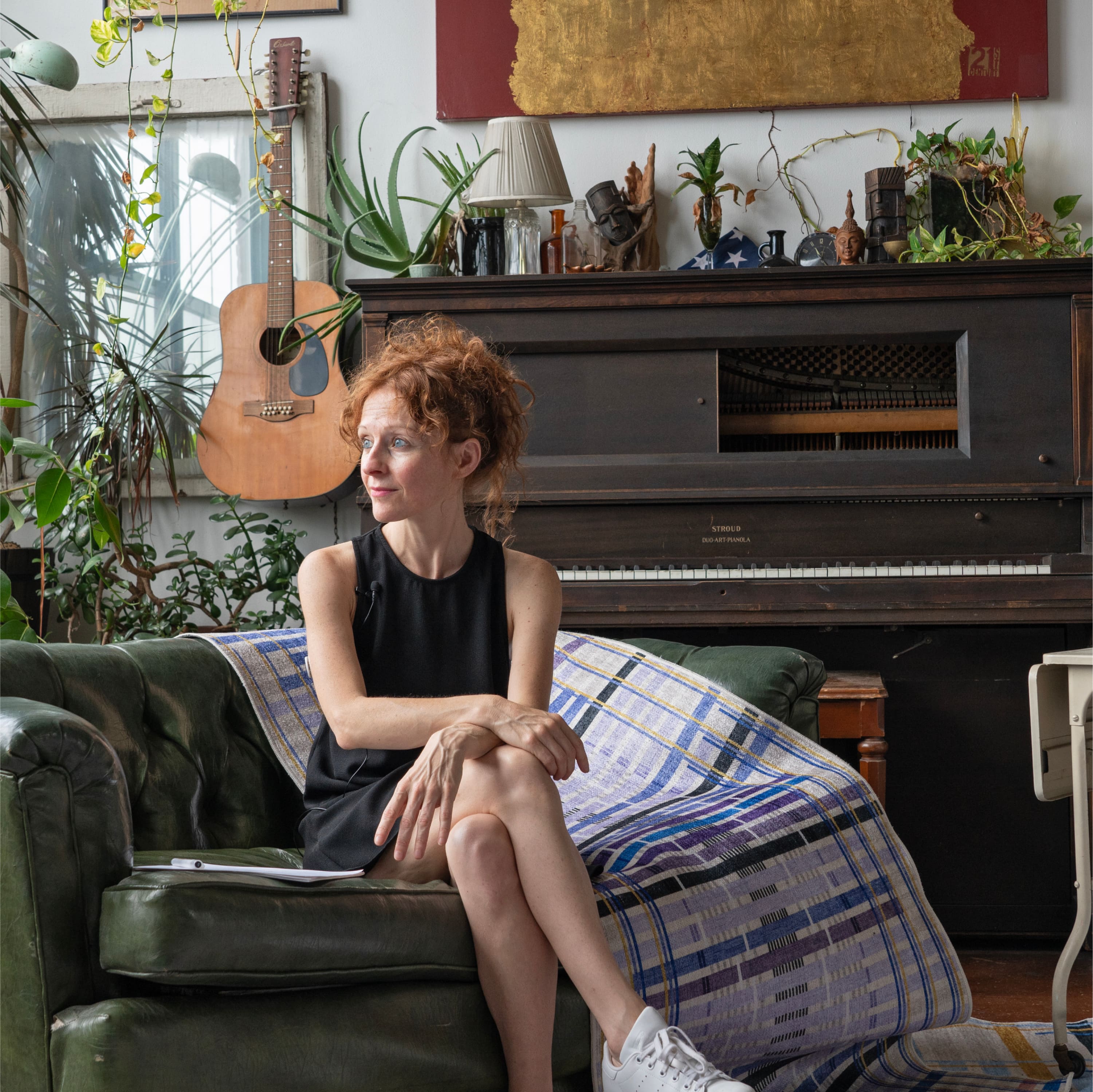 Data humanist Giorgia Lupi sitting on a green couch in a room with a piano and plants. Her Unraveling Stories rug is displayed next to her.