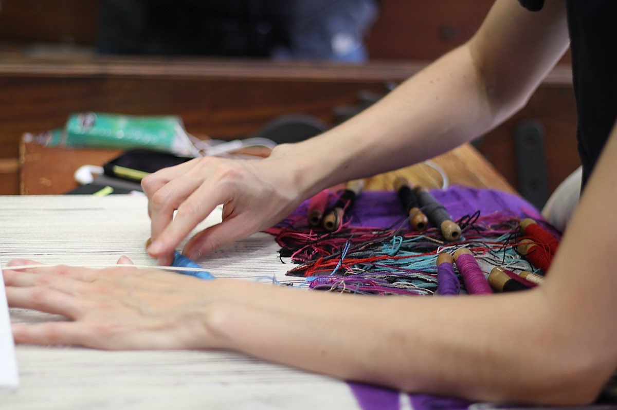 A weaver's hands on the warp of a low-warp loom in Aubusson, wooden bobbins of colored wool resting on the work.