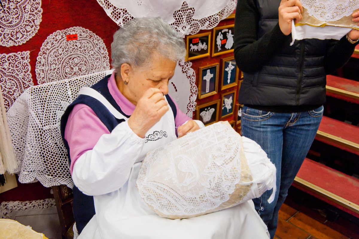 A lacemaker on Burano bent over her pillow working needle lace, finished merletto displayed on the wall behind her.