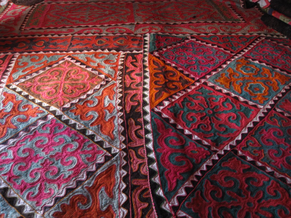 Shyrdak felt carpets layered inside a Kyrgyz yurt, their mosaic appliqué scrollwork filling the frame.