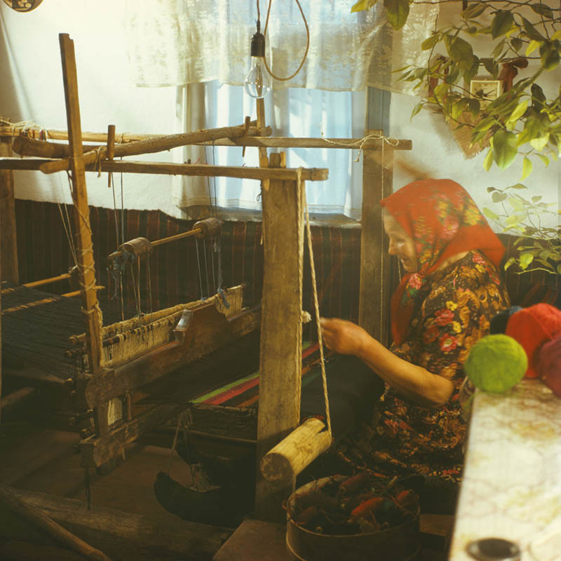 A Moldovan weaver in a flowered headscarf working at a traditional wooden loom, photographed in 1985.