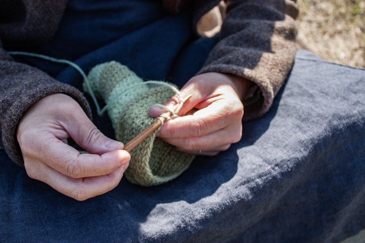 Hands drawing a wooden needle through green wool, a nålebinding sock in progress.