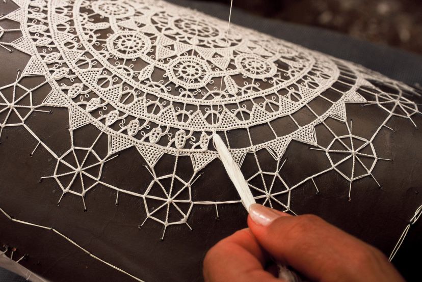 A lacemaker's hand guiding needle and thread over a half-finished Pag lace rosette pinned to a dark cushion.