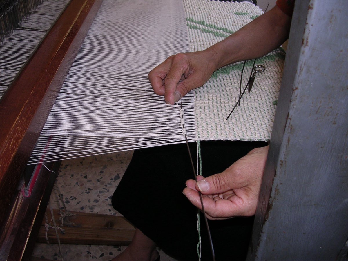 A weaver's hands at a loom in Ulassai, Sardinia, winding weft around the metal rod that raises the looped pibiones pile.