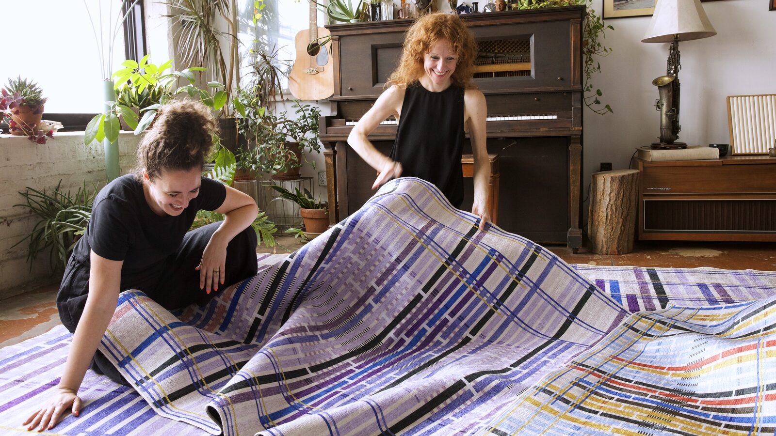Giorgia Lupi and a collaborator unrolling the Lavender colorway of Unraveling Stories over the Multi colorway, laughing, in a plant-filled room